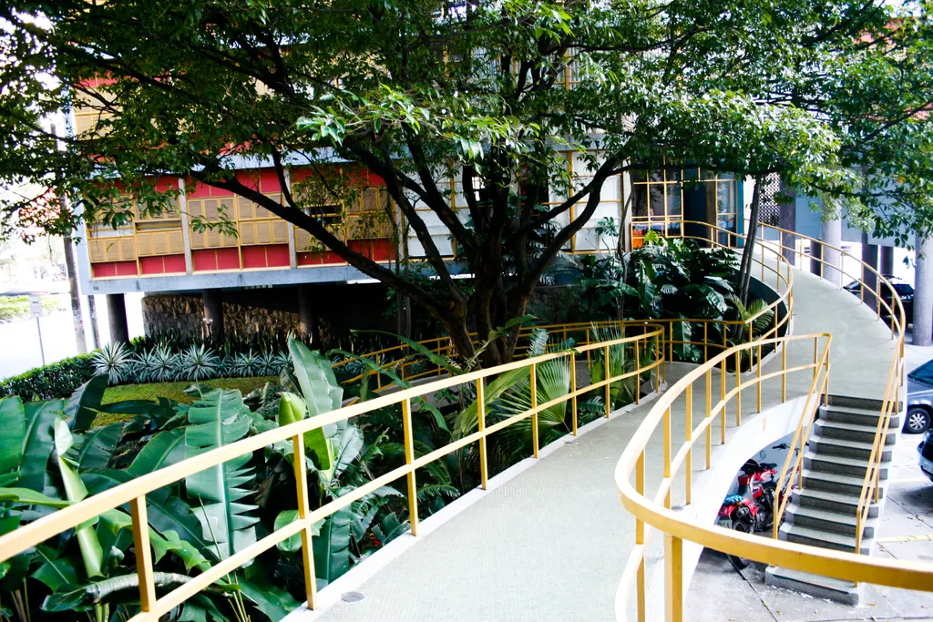 Rampa curva com guarda-corpo amarelo atravessando o jardim do Edifício Louveira, entre árvores e plantas tropicais
