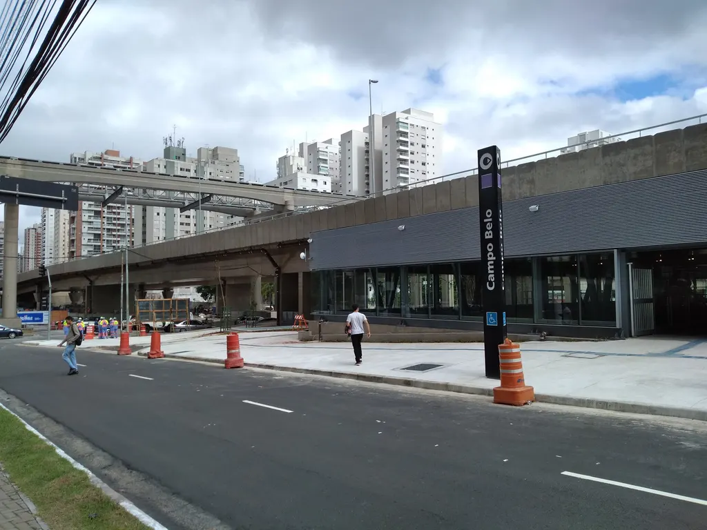 Entrada da estação Campo Belo, com totem do metrô na calçada e a via elevada do monotrilho ao fundo